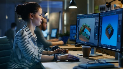 Eine Person sitzt an einem Tisch, auf dem zwei Bildschirme und eine Tastatur stehen. Auf den Bildschirmen sind Programme geöffnet. Die Person blickt auf die Bildschirme