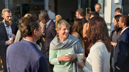 Mehrere Personen stehen in kleinen Gruppen im Freien und unterhalten sich. Manche Personen halten einen PappbecherHinter den Personen ist eine Glastür