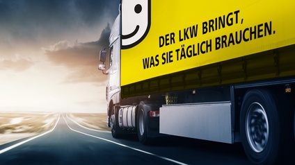 Fahrender Lkw mit Aufschrift „Der Lkw bringt, was Sie täglich brauchen“ auf Landstraße