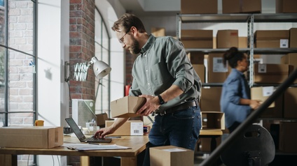 Personen arbeiten im Lagerraum am Laptop, bereiten Pakete für die Auslieferung vor und verpacken sie.