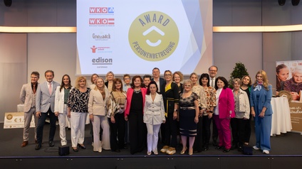 Eine Gruppe von rund 20 Personen steht nebeneinander auf einer Bühne und blickt in die Kamera. Im Hintergrund ist eine große Projektionsfläche mit einem runden gelben Logo und dem Text „AWARD PERSONENBETREUUNG“