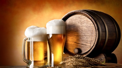 Auf einem Holztisch sind Getreideähren, zwei unterschiedliche Biergläser mit Bier sowie ein kleines Bierfass