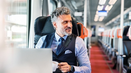 Bärtige Person mit grauen Haaren in Gillet im Portrait auf Einzelplatz in Zug sitzend, zur Seite blickend, hält Kaffeetasse in Hand, vor ihr verschwommen aufgeklappter Laptop, im Hintergrund roter Teppichboden und Sitzplätze