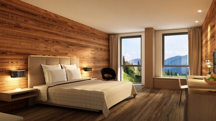 Hotelzimmer mit Wänden und Boden aus Holz, mittig ein beiges Doppelbett, im Hintergrund Fenster mit Blick auf Berge und Wald, Lichtstimmung goldig