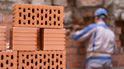 Im Vordergrund sind sechs Ziegelsteine übereinander gestapelt. Im Hintergrund ist der Rücken einer Person mit Helm, die direkt vor einer Mauer aus Ziegeln steht