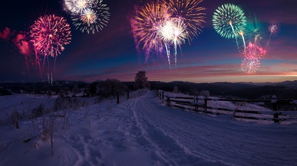 Eine verschneiter Weg bzw. Landschaft. Der Weg wird von einem Holzzaun begrenzt. Am dunklen Himmel ist ein buntes Feuerwerk