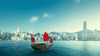 Auf einer Meeresbucht fährt ein zweistöckiges Holzschiff. Im Hintergrund ist die Skyline der Stadt Hongkong