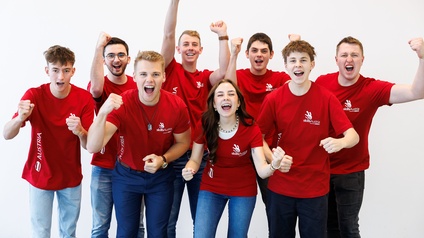 Porträt von acht jugendlichen Personen in roten Oberteilen, die nebeneinander vor einer weißen Wand stehen und jeweils jubeln