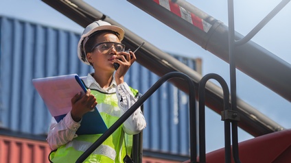 Eine Person mit Warnweste, Walkie-Talkie, einem Helm, Schutzbrille und Klemmbrett im Arm steht vor einem Stapel Container. Die Person spricht in das Walkie-Talkie.