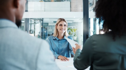 Ein an einem Tisch mit anderen Personen sitzende Person bekommt  Dokumente ausgehändigt.
