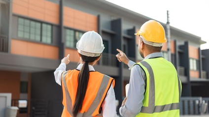 Rückansicht zweier Menschen in Warnwesten und Sicherheitshelmen die auf ein Gebäude zeigen