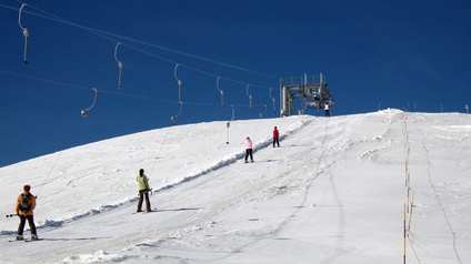 Rückenansicht von fünf versetzen Personen an einem schneebedeckten Hang in Skikleidung, Skischuhen und Ski, die sich an schrägen Stangen festhalten, die an einem Seil befestigt sind. 