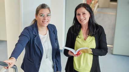 Zwei Personen stehen in einem Raum und blicken freudig in die Kamera, eine Person hält ein Buch in Händen