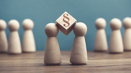 Auf einer hölzernen Oberfläche stehen im Vordergrund zwei kleine Holzfiguren, zwischen deren Köpfen ein Holzwürfel gehalten wird, auf dem ein Paragrafensymbol ist. Im Hintergrund stehen weitere Holzfiguren