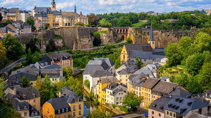 Blick über eine dicht bebaute Stadt mit historischen Gebäuden und grauen Dächern. Zwischen den Häusern verläuft ein schmaler Wasserlauf. Im mittleren Bildbereich stehen eine Kirche mit hohem, spitzem Turm und weitere Gebäude
