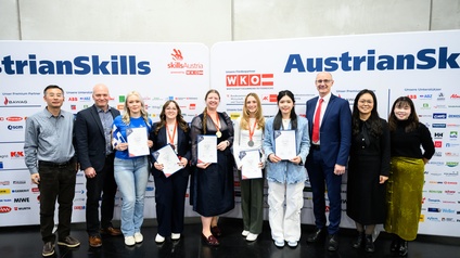 Gruppenfoto von zwölf Personen, die vor einer Plakatwand stehen, auf der verschiedene Logos von Firmen sind. Darüber steht AustrianSkills. Fünf der Personen halten eine Urkunde in die Kamera. Drei der Personen haben eine Medaille um den Hals
