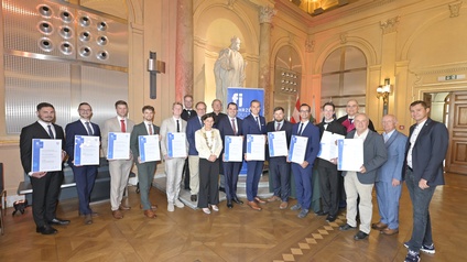 Porträt von vielen Personen in Anzügen, die nebeneinander in einem hohen Raum stehen. Die meisten der Personen halten jeweils eine große Urkunde