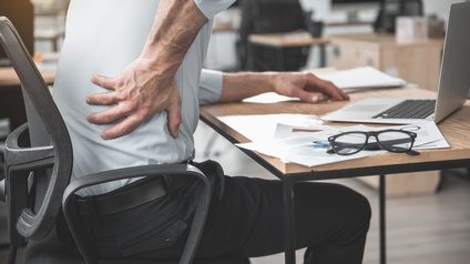 Arbeitnehmer sitzt bei der Arbeit am Tisch und hält sich wegen Rückenschmerzen den Rücken. Symbolbild für gesundheitliche Belastungen am Arbeitsplatz.
