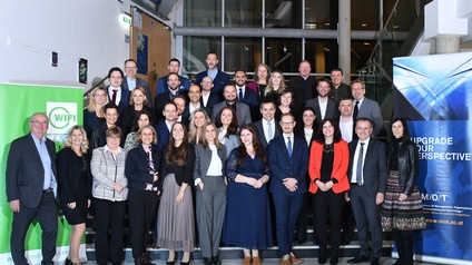 Gruppenfoto von vielen Personen in festlicher Kleidung, die hinter- und nebeneinander stehen. Links neben ihnen ist ein Banner, auf dem unter anderem WIFI steht