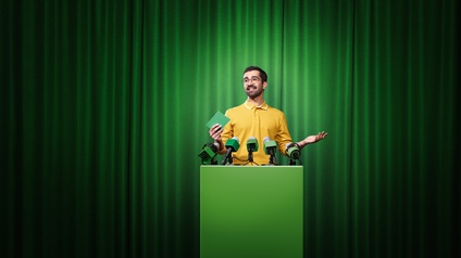 Person mit Brillen und gelbem Hemd steht an grünem Rednerpult mit Mikrofonen, im Hintergrund grüner Vorhang
