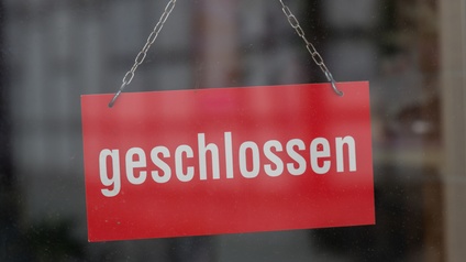 Vor einem grauen, verschwommenen Hintergrund sieht man ein Schild in der Bildmitte, das an einer Metallkette hängt. Das Schild ist rot und trägt die Inschrift 