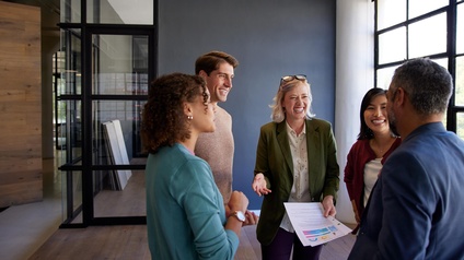 In einem Büroraum stehen fünf Personen in einem Kreis und unterhalten sich. Die Personen lachen teilweise. Eine der Personen hält einen Zettel mit verschiedenen Diagrammen