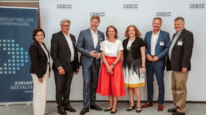 Gruppenfoto mit sieben Personen in Business-Kleidung. Die Personen stehen nebeneinander vor einer weißen Hintergrundwand mit mehrfach platziertem WKÖ-Logo. Links im Bild befindet sich ein blaues Roll-up der Industriellenvereinigung mit dem teilweise lesbaren Text „Industriellen Vereinigung“ sowie „Zukunft gestalten“. Alle Personen tragen Namensschilder