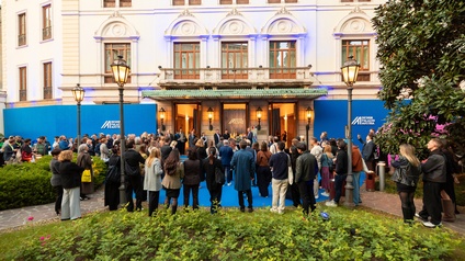 Vor einem historischen Gebäude versammelt sich eine größere Personengruppe auf einem blauen Teppich. Der Eingangsbereich ist geöffnet und beleuchtet. Über dem Eingang befindet sich die Beschriftung „Design Palazzo Austria“