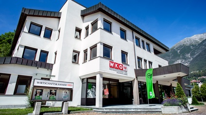 Außenansicht der Bezirksstelle Imst der Wirtschaftskammer Landeck. Das moderne, dreigeschossige Gebäude mit weißer Fassade und großen Fenstern trägt das WKO-Logo über dem Haupteingang. Vor dem Eingang stehen eine grüne WIFI-Fahne und eine Informationstafel der Wirtschaftskammer. Im Hintergrund sind Berge und ein klarer blauer Himmel zu sehen.