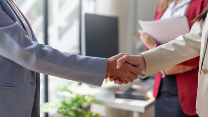 Seitliche Aufnahme zweier Personen in Sakkos, die sich gegenüber stehen und die Hände schütteln. Neben ihnen steht eine weitere Person, die einen Stapel Zettel hält