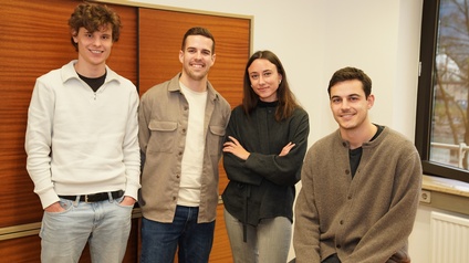 Vier junge Erwachsene stehen bzw. sitzen in einem Büroraum und lächeln in die Kamera. Drei Personen stehen nebeneinander, eine Person sitzt rechts auf einer Tischkante. Der Raum hat weiße Wände, ein großes Fenster und Holzschränke im Hintergrund.