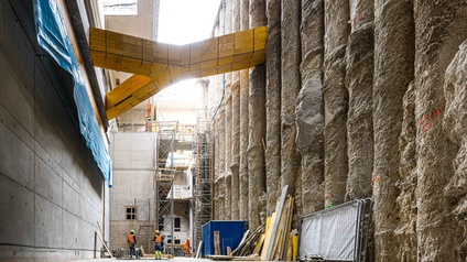 Zwei Personen mit Helm und Schutzweste arbeiten auf einer Baustelle. Man sieht Gerüste, Bretter und Dämmmaterial. Auf die Baustelle fällt Tageslicht 