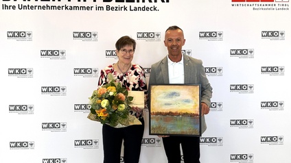 Zwei Personen stehen vor einer WKO-Tirol-Fotowand im Bezirk Landeck. Eine Frau hält einen Blumenstrauß und ein Kuvert, ein Mann präsentiert ein gerahmtes Landschaftsbild. Beide schauen in die Kamera.