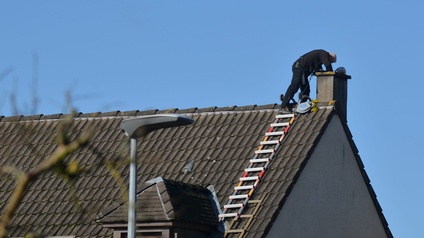 Eine Person, komplett in schwarz gekleidet, steht auf der Spitze eines Dachs, direkt neben einem Kamin, und beugt sich über diesen. Die Hände der Person sind im Kamin. Unter der Person ist am Schrägdach eine Leiter befestigt