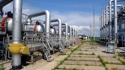 Metallische Gastanks mit Röhren gegenüberliegend , in der Mitte betonierter, von Pflanzen durchbrochener Weg unter blauem Himmel mit Wolken