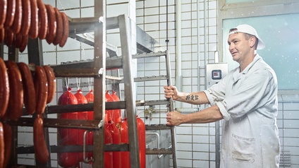 Person in weißer Arbeitskleidung in Fleischereibetrieb zieht Metallgestell mit verpackten Wurstwaren