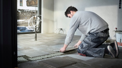 Ein Mann verlegt graue Bodenfliesen. Im Vordergrund Stapel noch zu legender Fliesen.