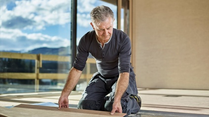 Person in grauer Arbeitsmontur kniet am Boden und verlegt Holzbodenbretter, im Hintergrund große Fensterfläche mit Blick auf blauen wolkendurchzogenen Himmel