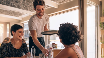 Zwei Personen sitzen sich an einem Tisch in einem Lokal gegenüber. Neben ihnen steht eine Person mit kurzen Haaren, Bart und Tablett, die eine Flasche Wasser vor eine der Personen stellt und lächelt