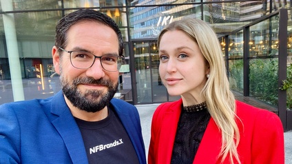 Porträt von zwei nebeneinander stehenden Personen vor einem Gebäude auf einem asphaltierten Platz. Die linke Person hat kurze, dunkle Haare und trägt Brille. Die rechte Person hat offene, helle Haare und trägt ein dunkles Oberteil
