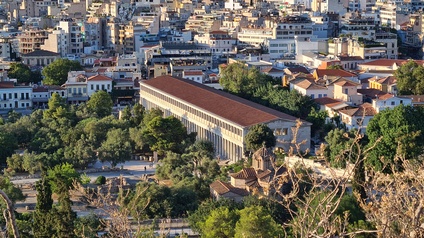 Das Foto zeigt ein langgestrecktes, rechteckiges Gebäude mit vielen gleichmäßigen Säulen entlang der Längsseite und einem roten Satteldach. Das Gebäude liegt inmitten einer dicht bebauten Stadt mit mehrstöckigen Häusern in unterschiedlichen Farben und Bauweisen. Im Vordergrund befinden sich Bäume und Grünflächen