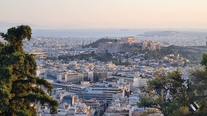 Panoramaaufnahme der Stadt Athen mit den verschiedenen Gebäuden sowie der Sehenswürdigkeit Akropolis. Im Hintergrund ist das Meer