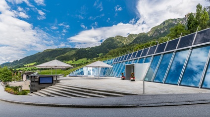 Panoramablick auf den Vorplatz des Congress Centrum Alpbach an einem sonnigen Tag. Das moderne Gebäude mit großen, schrägen Glasfassaden und Solarpaneelen liegt vor einer Bergkulisse mit grünen Wäldern und Wiesen. Auf dem Platz stehen zwei große Sonnenschirme, Blumenarrangements und Sitzgelegenheiten.