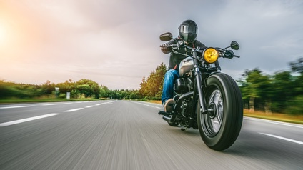 Frontalansicht einer Person auf einem Motorrad. Das Motorrad ist auf einer asphaltierten Straße. Die Person trägt einen Helm und Handschuhe. Links und rechts von der Straße sind grüne Bäume