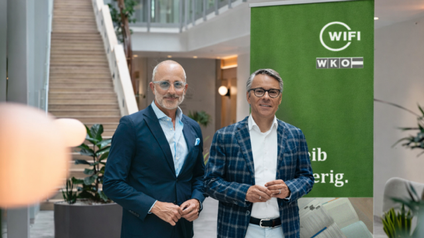 Porträt von zwei Personen in Sakkos sowie mit grauen Haaren und Brillen, die nebeneinander in einem Foyer stehen. Hinter ihnen ist ein grüner Banner, auf dem WKO und WIFI steht