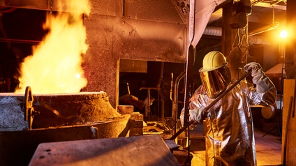 Person in feuerabweisender Schutzkleidung mit Helm und Gesichtsvisier hält Stange, daneben großer Metallbehälter aus dem Flammen emporsteigen