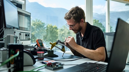 Ein junger Mann sitzt im Büro vor einem Computer, er lötet an einem kleinen Gerät. Im Vordergrund ein offener Laptop