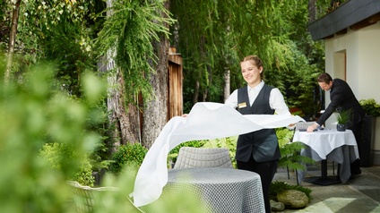 Lächelnde Person mit Zopf in grauem Gilet und weißem Hemd breitet weißes Tischtuch über Tisch in Garten, im Hintergrund weitere Person, die anderen Tisch eindeckt, ringsum Grün der Pflanzen