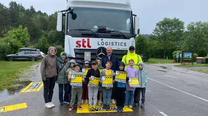 Gruppe von Schulkindern und Begleitpersonen steht vor einem weißen Lkw mit „stl logistics“-Logo; mehrere Kinder halten gelbe Warnmaterialien mit der Aufschrift „LKW FRIENDS“; Aufnahme im Freien auf einem Parkplatz.
