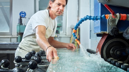 Ein Mann schleift eine frisch geschnittene Glasscheibe an der Maschine. Gegen die Staubbildung wird Wasser darauf gespritzt.